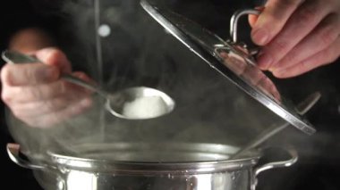 Elleri bir kaşıktan ocakta buharlaşan bir tencereye tuz döküyor. Tencere metalden yapılma ve mutfak aktiviteleriyle dolu..