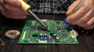 Bir insan atölye ayarında lehim ve lehim kablosu kullanarak yeşil devre kartının parçalarını lehimliyor. Cihazlar ve malzemeler yakında.