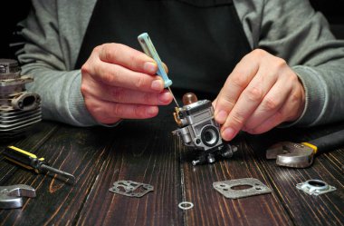 Bir insan atölyede tornavida kullanarak bir budama karbüratörünü tamir etmeye çalışır. Çeşitli aletler ve parçalar ahşap masaya yayılmış..