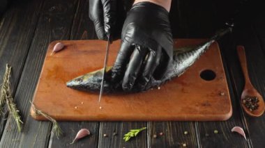 Siyah eldivenli biri tahta kesme tahtasındaki uskumru balığını dilimliyor. Sarımsak, otlar ve baharatlı ahşap bir kaşık mutfak masasındaki balığın etrafını sarar..