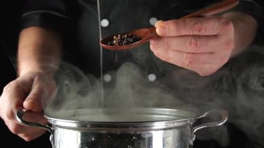 Bir aşçı sobadaki bir kaba baharat eklemek için tahta kaşık kullanır. Şef yemek pişirmeye odaklanırken tencereden buhar yükseliyor. Sahne, detaylara dikkat eden yoğun bir mutfağı gösteriyor..