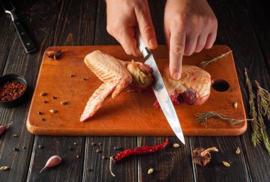 Elleri, tahta bir kesme tahtasıyla çiğ et keserken bıçak tutuyor. Gündüz vakti mutfak alanında çeşitli baharatlar ve otlar tablonun etrafını sarar..