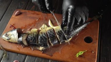 Bir aşçı büyük bir uskumru balığını tahta bir tahtanın üzerinde parçalara ayırıyor. Sahnede mutfak tezgahının yanında çeşitli mutfak gereçleri ve malzemeler var..