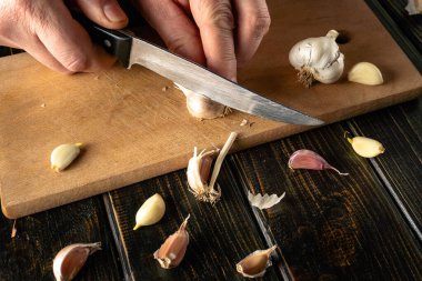 Eller, ahşap bir tahtanın üzerindeki sarımsak ampulünü kesmek için bıçak kullanıyor. Tahtanın etrafına birkaç sarımsak parçası saçılmış. Işık kalitesi gündüz olduğunu gösteriyor..