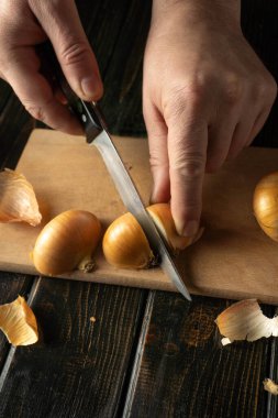 Bir insan mutfaktaki tahta bir tahtada soğan kesmek için bıçak kullanır. Sahne, ellerin masaya yakın soğan kabuklarıyla göreve odaklandığını gösteriyor..