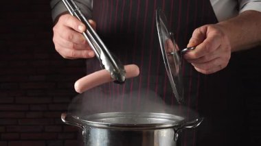 Çizgili önlüklü bir şef sıcak sosisi maşayla sıcak bir tavaya yerleştirirken diğer elinde kapağı tutuyor ve mutfak ortamındaki pişirme sürecini vurguluyor..