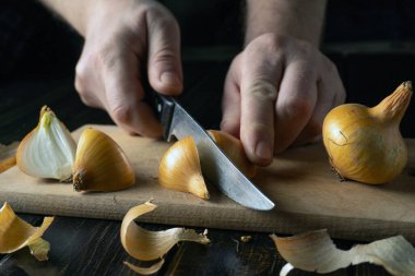 Bir insan mutfaktaki tahta kesme tahtasındaki soğanları dilimlemek için bıçak kullanır. Arka plan karanlık bir ortam gösteriyor. Kabukları etrafa saçılmış..