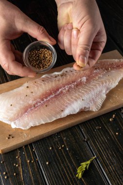 Tahta kesme tahtasına yerleştirilmiş taze balık filetosuna baharat serpiyor. Bu sahne bir mutfakta geçiyor, yemek pişirme aktivitelerini ön plana çıkarıyor..