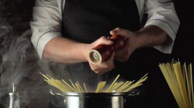 Siyah önlüklü bir şef paslanmaz çelik bir tencerede pişmemiş spagetti ve mutfak elementleriyle çevrili haşlanmış makarnanın üzerine biber öğütür ve kırsal bir mutfakta lezzetli bir yemek yaratır.