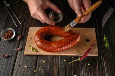 Bir insan, ahşap kesme tahtasındaki bir sosise yağ sürer. Baharat ve sarımsak tahtanın etrafını sarmış. Mutfak aletleri yakında. Dekorasyonda bir ev mutfağında yemek hazırlığı var..