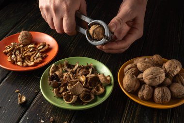 Bir insan ceviz kırmak için fındık kırıcı kullanır. Açılmış fındıklar yeşil bir tabağa yerleştirilirken, kabuklar ve cevizler turuncu ve kahverengi tabaklara konur..