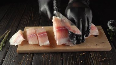 Siyah eldivenli şef, ahşap kesme tahtasındaki taze balık filetolarını ustalıkla dilimliyor, etrafa saçılmış otlar ve baharatlarla, mutfak ortamındaki aşçılık hazırlama sürecini vurguluyor.
