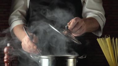 Siyah önlüklü şef buharlı tencerenin kapağını kaldırıyor, içinde makarna olduğunu gösteriyor, mutfak tezgahında spagetti ve biber değirmeni düzenleniyor, mutfak atmosferi yaratıyor.