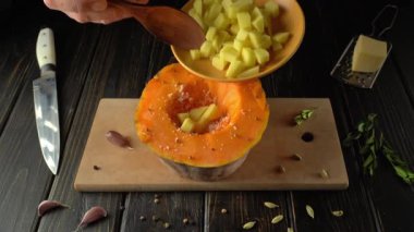 Mutfak ortamındaki bir insan içi oyulmuş bir balkabağına doğranmış patates eklemek için tahta kaşık kullanır. Mutfak aletleri, baharatlar ve ahşap tezgahtaki taze malzemelerle çevrili.