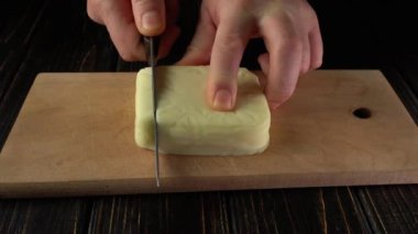 Elleri ahşap bir kesme tahtasının üzerine yerleştirilmiş, bir parça peyniri küçük parçalara ayırıyor, iyi aydınlatılmış bir mutfak ortamında tereyağının kremsi dokusunu ve pürüzsüz yüzeyini ortaya çıkarıyor.