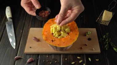 Eller, ahşap kesim tahtasındaki oyuk bir balkabağına dikkatlice baharat ve peynir ekliyor. Yanında da mutfak bıçağı ve sarımsak karanfilleri var.