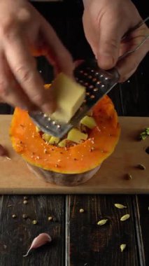 Şef, ahşap kesim tahtasındaki baharat ve otlarla dolu bir balkabağının üzerinde peynir rendeliyor ve kırsal bir mutfak ortamındaki malzemelerin karmaşık detaylarını gözler önüne seriyor.