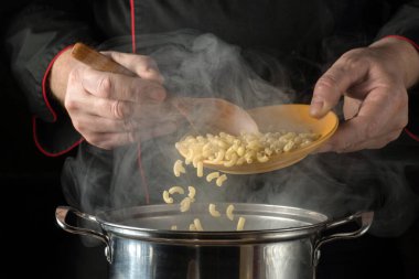 Bir şef mutfakta makarna hazırlıyor. Sarı tabaktan tencereye makarna aktarmak için tahta kaşık kullanıyor. Buhar yükseliyor, yemek pişirmenin sıcaklığını gösteriyor..