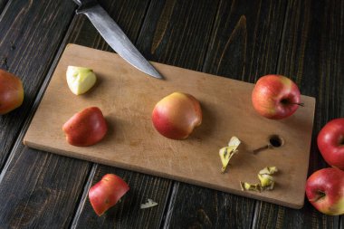 Tahta bir kesme tahtası karanlık bir masanın üzerine yerleştirilir. Kırmızı elmalar parçalara ayrılırken yakınlarda bir bıçak duruyor. Tahtada bazı elma kırıntıları var, yemek hazırlıklarını gösteriyor..