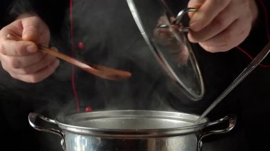 Şefin elleri tencerenin kapağını kaldırırken ahşap kaşıkla içindekileri karıştırıyor, karanlık bir mutfakta buhar yükseliyor, yemek pişirme sürecini ve mutfak atmosferini gösteriyor.