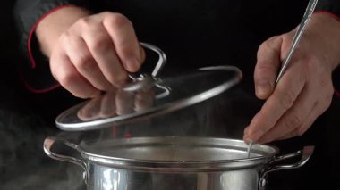 Şef tencerenin kapağını tek eliyle kaldırırken kepçeyle buharda pişmiş tahılları topluyor ve mutfak ortamındaki bezelyeleri gösteriyor.