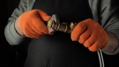Gri gömlekli ve turuncu eldivenli şahıs, loş aydınlatılmış bir atölye ortamında el konumu ve araç etkileşimine odaklanarak, bir ingiliz anahtarı kullanarak metal tesisatlara bağlanır.