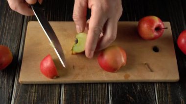 Ellerinde bir bıçakla ahşap kesme tahtasındaki kırmızı elmaları kesiyorlar. Elma parçaları etrafa saçılmış ve elmalar iyi aydınlatılmış bir mutfak ortamında görülebiliyor.