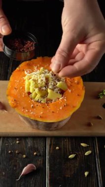 Eller dikkatlice, ahşap kesim tahtasındaki peynir ve otlarla dolu bir balkabağının üzerine baharat serpiyor, sarımsak ve baharatlar çevrede görülebiliyor, yemek pişirme sahnesini güçlendiriyor.