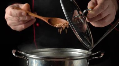 Siyah üniformalı bir şef buğday buğdayını ahşap bir kaşıkla karıştırır, kapağı kaldırır, loş bir mutfak ortamında kasıtlı pişirme sürecini gösterir..