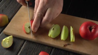 Elleri kırmızı ve yeşil elmaları dilimleyen bir bıçakla tahta kesme tahtasına yerleştirilmiş. Mutfak ortamında sürekli kesme ve elma parçalarının düzenlenmesini gösteriyor.