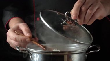 Siyah üniformalı bir şef paslanmaz çelik bir tencerenin kapağını kaldırır. Koyu bir mutfak ortamında buhar dalgalanırken tahta kaşıkla pirinç eklemeye hazırlanır.