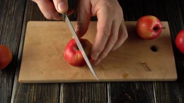 Ellerin yakın plan çekimleri, tahta kesme tahtasındaki kırmızı bir elmayı kesmek için bıçak kullanırken, diğer elmalar olay yerinin etrafına yerleştirilirken, iç meyve ve çekirdekleri ortaya çıkarıyor.