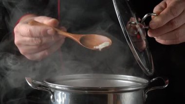 Siyah üniformalı şef tencerenin kapağını kaldırırken tahta kaşıkla pirinç ekliyor, tencereden buhar yükseliyor, modern bir mutfak ortamında pişirme sürecini vurguluyor.
