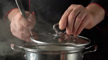 Şefin elleri paslanmaz çelik bir tencerenin kapağını kaldırırken, buhar yükselirken malzeme ya da darı eklemeye hazırlanırken, profesyonel bir mutfak ortamında odaklanmış bir pişirme süreci sergilerken görülüyor.