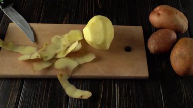 Mutfak ortamında soyulmamış patateslerin yanında soyulmuş patates ve soyulmuş patateslerin yer aldığı bir bıçakla ahşap kesim tahtasının üzerinde sergilenen patates soyma işlemi.