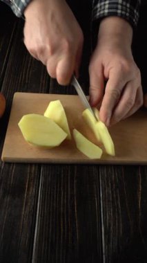 Damalı gömlekli bir insanın elleri tahta bir kesme tahtasının üzerinde taze patatesleri dilimliyor. Birden fazla patates parçası görülebiliyor. Mutfakta devam eden yemek hazırlama sürecini vurguluyor.