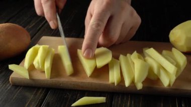 Eller ustalıkla taze patatesleri ince şeritlere ayırıyor, ahşap bir kesim tahtasının üzerinde, bütün bir patates ve sahneyi güçlendiren koyu ahşap bir yüzeye sahip kırsal bir mutfak arka planı var.