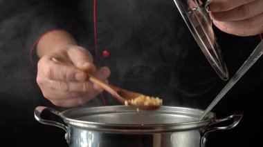 Bir şef mutfakta bir yemek hazırlar. Buhar yükselirken bir tencereyi tahta kaşıkla karıştırıyor. Atmosfer mutfak sesleri ve lezzetli aromalarla dolu..