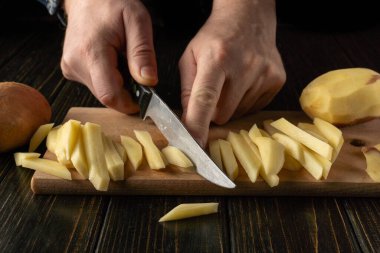 Eller, tahta bir kesme tahtasıyla çiğ patatesleri ince ince dilimlerken görülüyor. Başka bir patates yakınlarda dinlenirken bıçak kesmek için kullanılır. Ayarlar bir mutfak sahnesi gösteriyor..