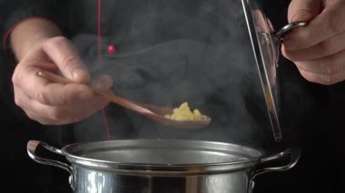 Siyah üniformalı şef, ahşap kaşıkla makarna eklemek için bir tencere kapağı açar. Paslanmaz çelik tencereden buhar sızar ve pişirme aromalarıyla dolu sıcak bir mutfak ortamı yaratır.