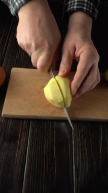 Kareli gömlekli bir insanın elleri, patatesin mutfak ortamında parçalara ayrılması gibi kesme eylemini gösteren tahta kesme tahtasındaki patatesi dilimliyor.