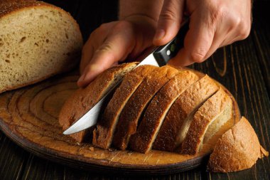 Elleri bir somun kahverengi ekmeği tahta kesme tahtasıyla kesiyor. Mutfağa doğal ışık geliyor ve ekmek taze..