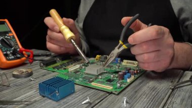 Teknisyen, iyi aydınlatılmış bir atölyedeki ahşap tezgahta çeşitli alet ve parçalarla çevrili lehim makinesi ve penseyle elektronik devre kartının onarımına odaklanıyor.