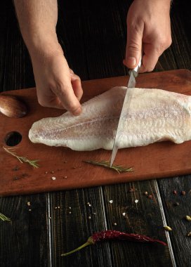 Mutfakta, bir insan ahşap bir tahtada balık filetosu keser. Taze bitkiler ve baharatlar yakına konur. Ayarlar yemek hazırlıklarını gösteriyor..