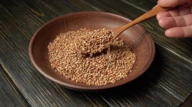 Bir el, karabuğday dolu ahşap bir kaşığı kaldırır. Kahverengi bir kâse kahverengi bir ahşap masada uzanır. Tohumların dokusunu ve kâsenin doğal görünümünü vurgular..