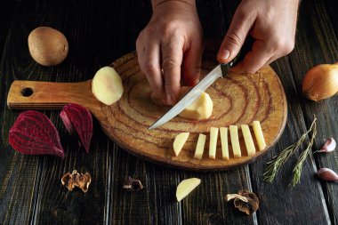 Eller, tahta kesme tahtasındaki patates ve soğanları dilimlemek için bıçakla çalışır. Diğer sebzeler kurulun etrafına dizilir. Sahne yemek hazırlıklarını gösteriyor..