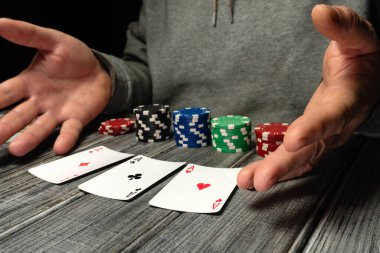 Masadaki bir kart oyununda üç iskambil kartı kazanan elleri gösteriyor. Farklı renklerde poker parçaları etrafa saçılmış. Oyun açılırken ellerinizi kaldırın..
