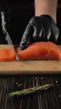 Siyah eldivenli şef, ahşap kesim tahtasında taze somon filetosunu ustalıkla dilimliyor. Etrafı otlar ve baharatlarla çevrili. Mutfak masasında, mutfak hazırlama teknikleri sergiliyor.