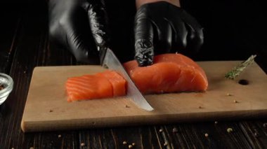 Siyah eldivenli şef ahşap kesim tahtasındaki taze somon filetosunu ustalıkla dilimliyor. Bitkiler ve baharatlar siyah ahşap masaya saçılmış ve mutfak sahnesi yaratıyor.