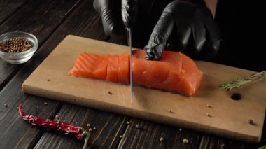 Siyah eldivenli şef, ahşap bir kesim tahtasında taze somon filetosunu ustalıkla dilimliyor. Etrafı otlar ve baharatlarla kaplı, ahşap bir masada canlı bir mutfak sahnesi yaratıyor.